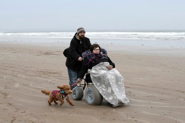A lady in a wheelchair accompanied by her partner and dog enjoy an amble along a sandy beach